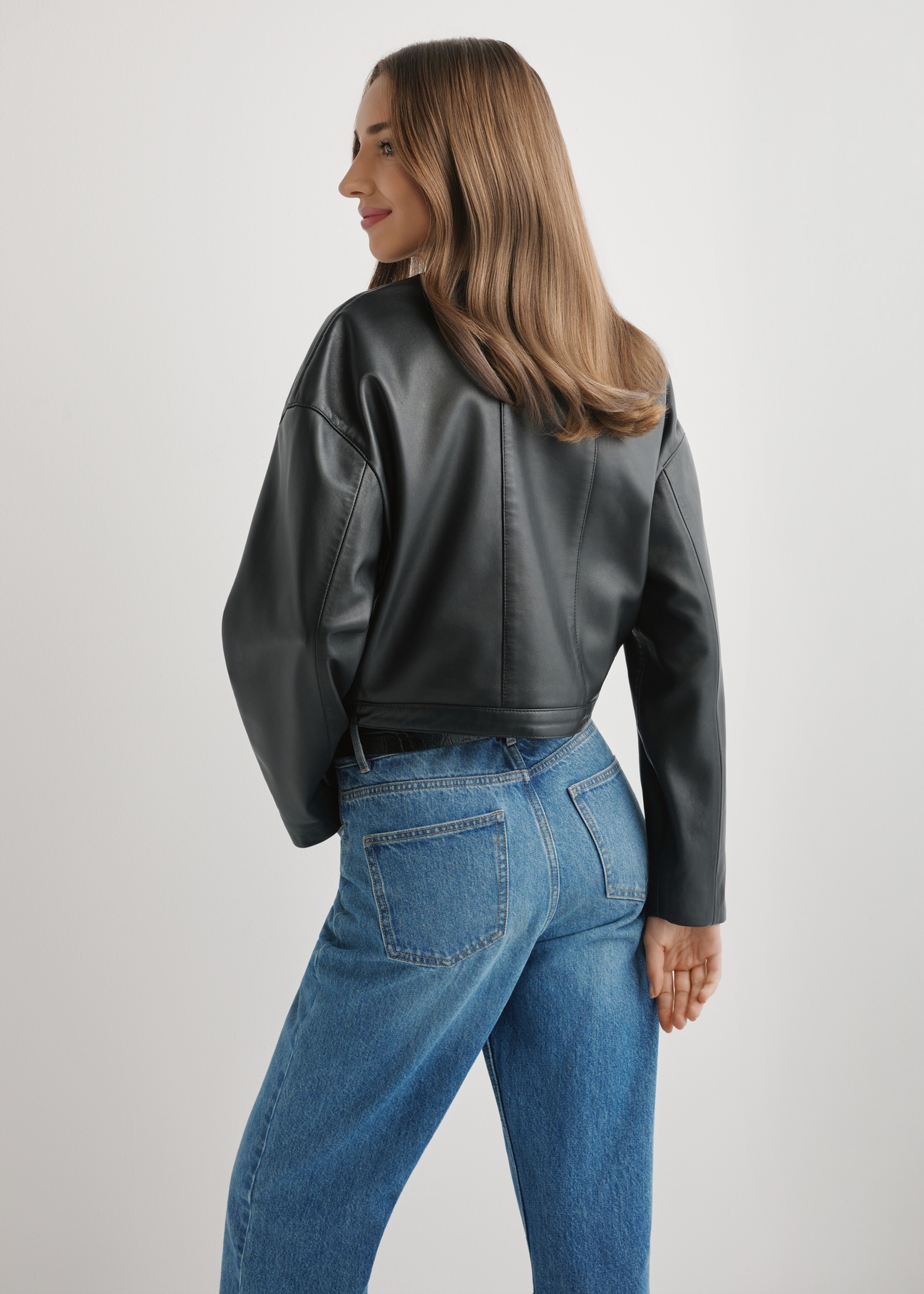 Krótka czarna kurtka skórzana damska KURDS-0597-99(W26), widok produktu z tyłu na modelce. Widoczna połowa sylwetki.
