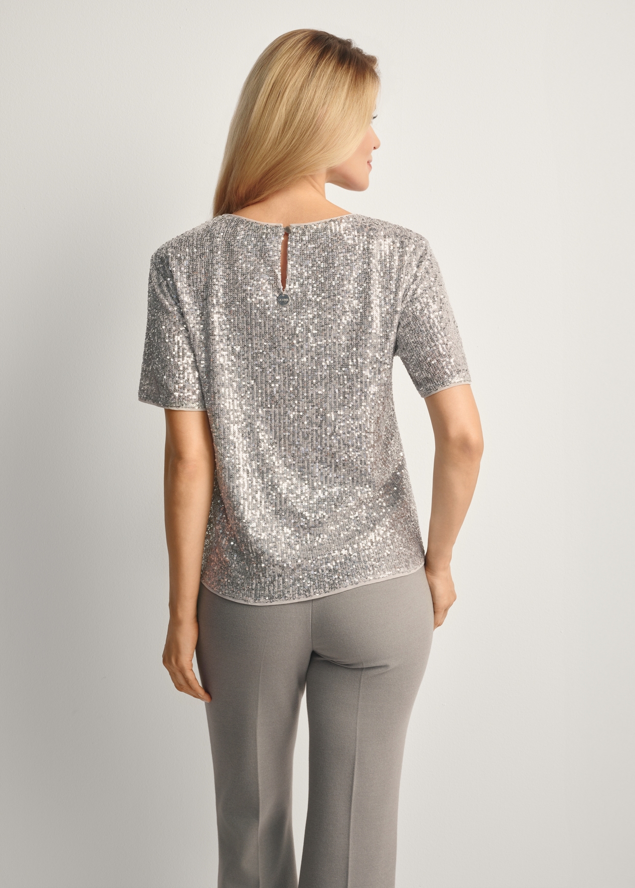 Srebrna cekinowa bluzka damska BLUDT-0199-92(Z25), widok produktu tyłem na modelce, widoczna połowa sylwetki.