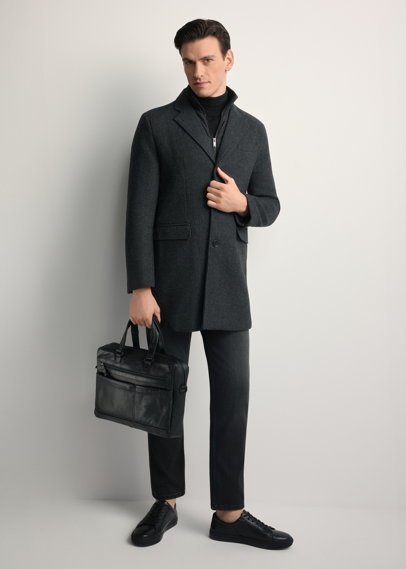 Czarna skórzana teczka męska TORMS-0415A-99(Z25) - widok produktu po skosie, trzymanego przez modela w ręku, na białym tle. Widoczna cała sylwetka. 