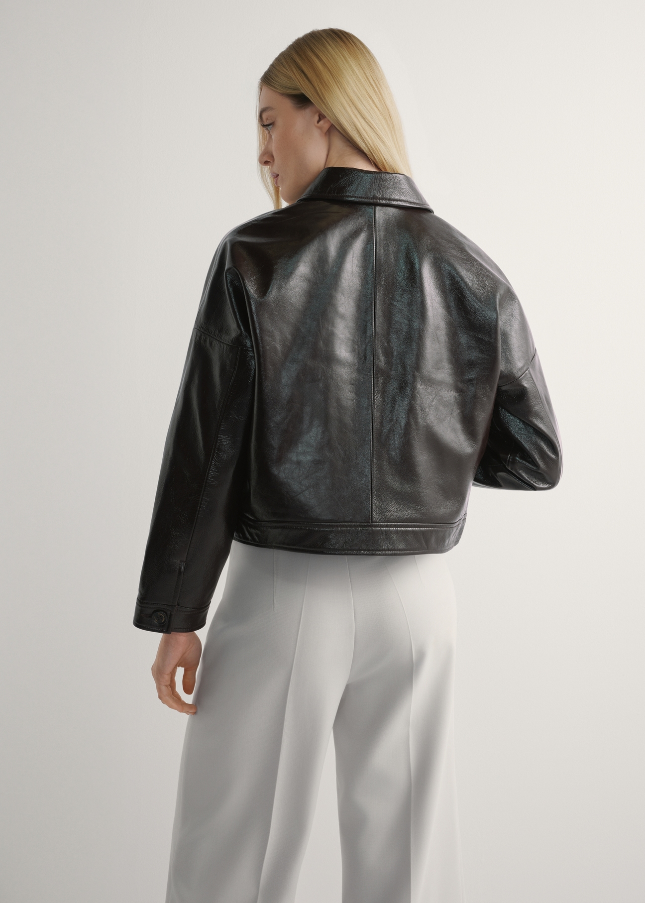 Brązowa skórzana krótka kurtka damska KURDS-0593-1I(W26) - widok produktu z tyłu, na modelce, na jasnym tle. Widoczna sylwetka do kolan. 