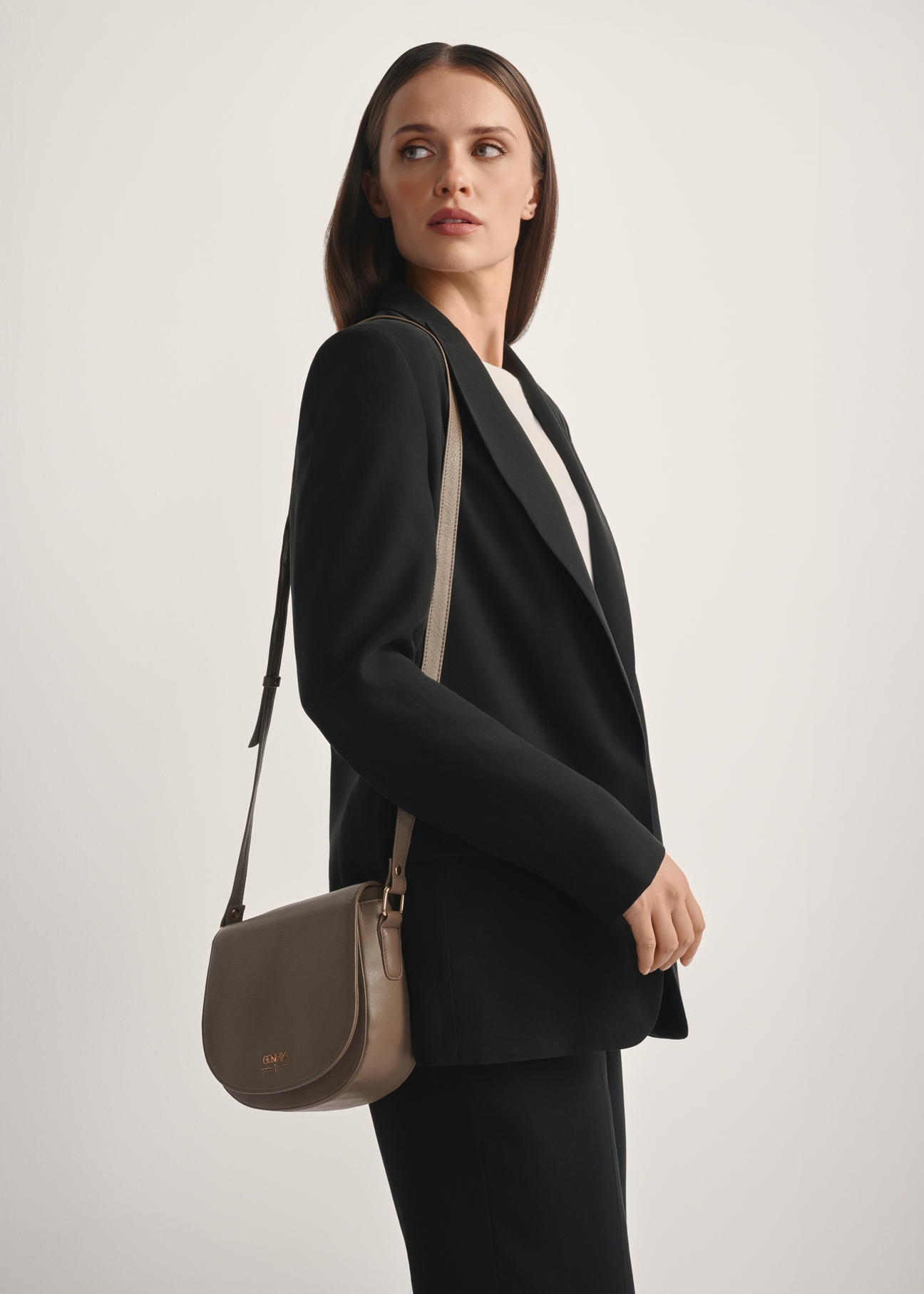 Ciemnobeżowa skórzana półokrągła torebka listonoszka TORES-0972F-1K(W26), widok produktu skosem na modelce, widoczna połowa sylwetki.