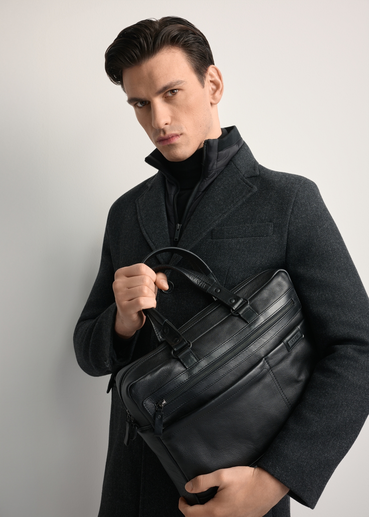 Duża czarna skórzana teczka męska TORMS-0416A-99(Z25) - widok produktu z przodu, trzymanego przez modela w ręku, na białym tle. Widoczna połowa sylwetki. 