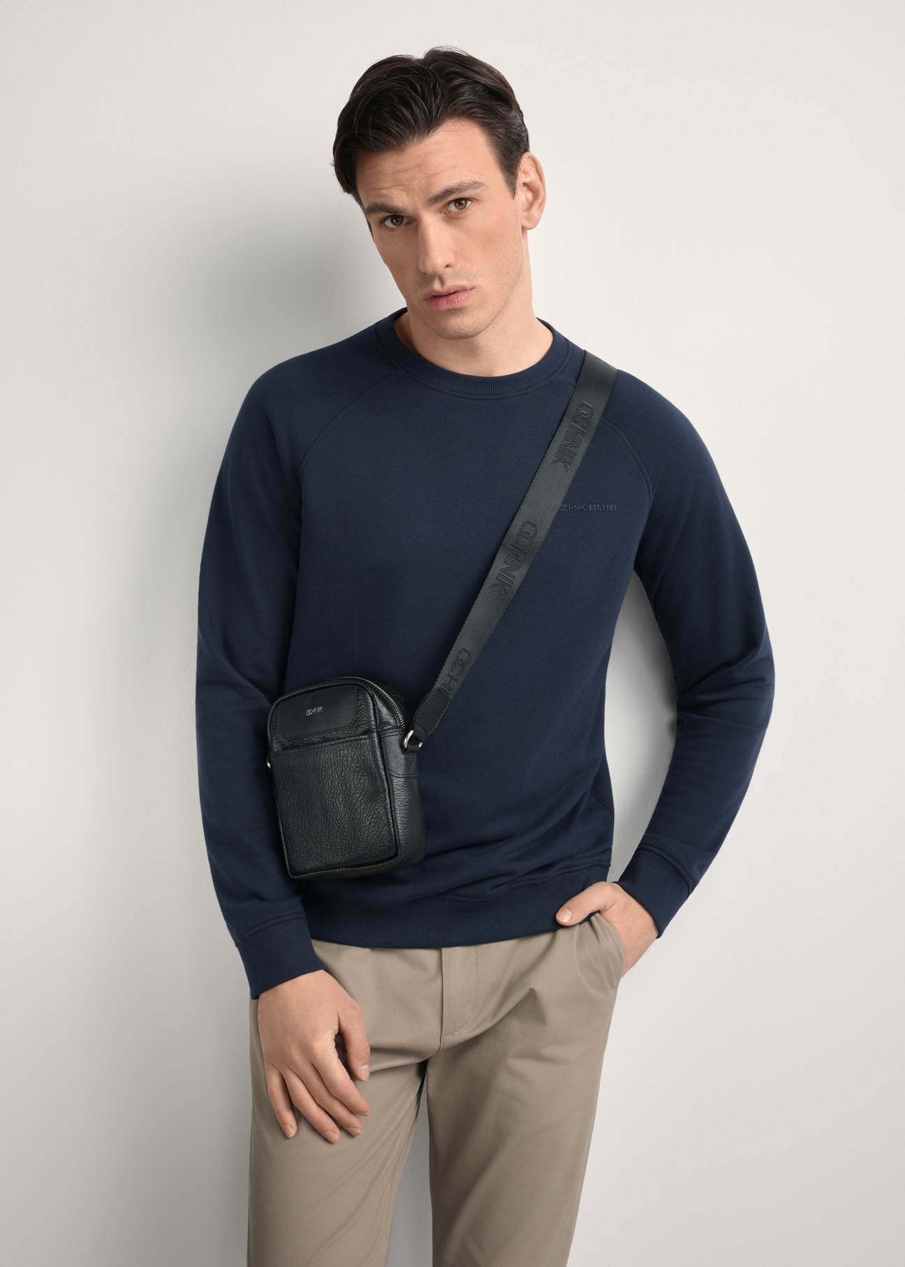 Czarna skórzana saszetka męska z logo TORMS-0296A-99(Z25) - widok produktu po skosie, na modelu, na białym tle. Widoczna sylwetka do kolan.