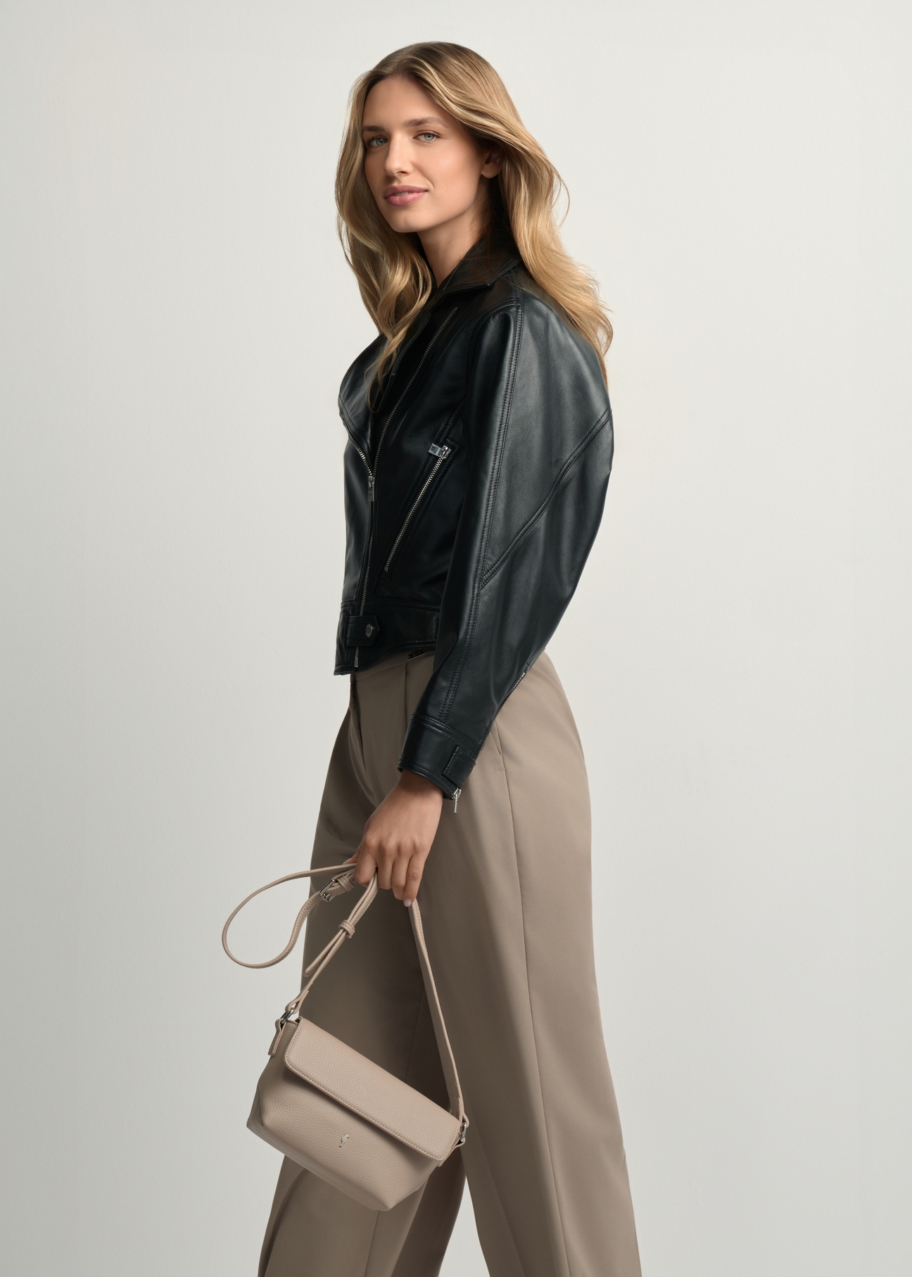 Beżowa mała torebka damska na ramię TOREC-0916-1A(W26)- widok produktu z przodu, trzymanego przez modelkę w ręku, na jasnym tle. Widoczna sylwetka do kolana.