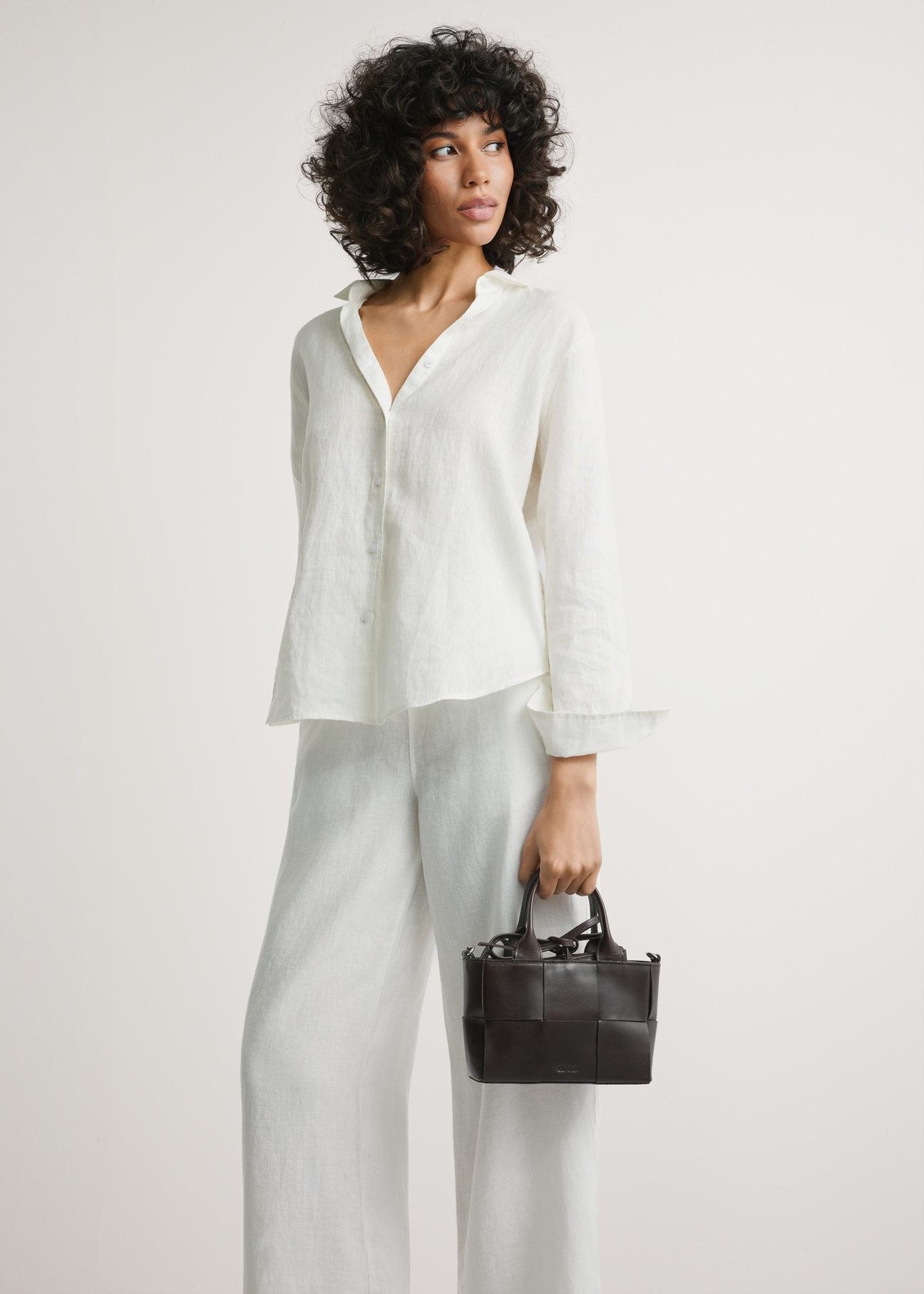 Ciemnobrązowa mała skórzana torebka damska TORES-1298-1H(W26) - widok produktu, trzymanego przez modelkę w ręku, na jasnym tle. Widoczna sylwetka do kolan. 