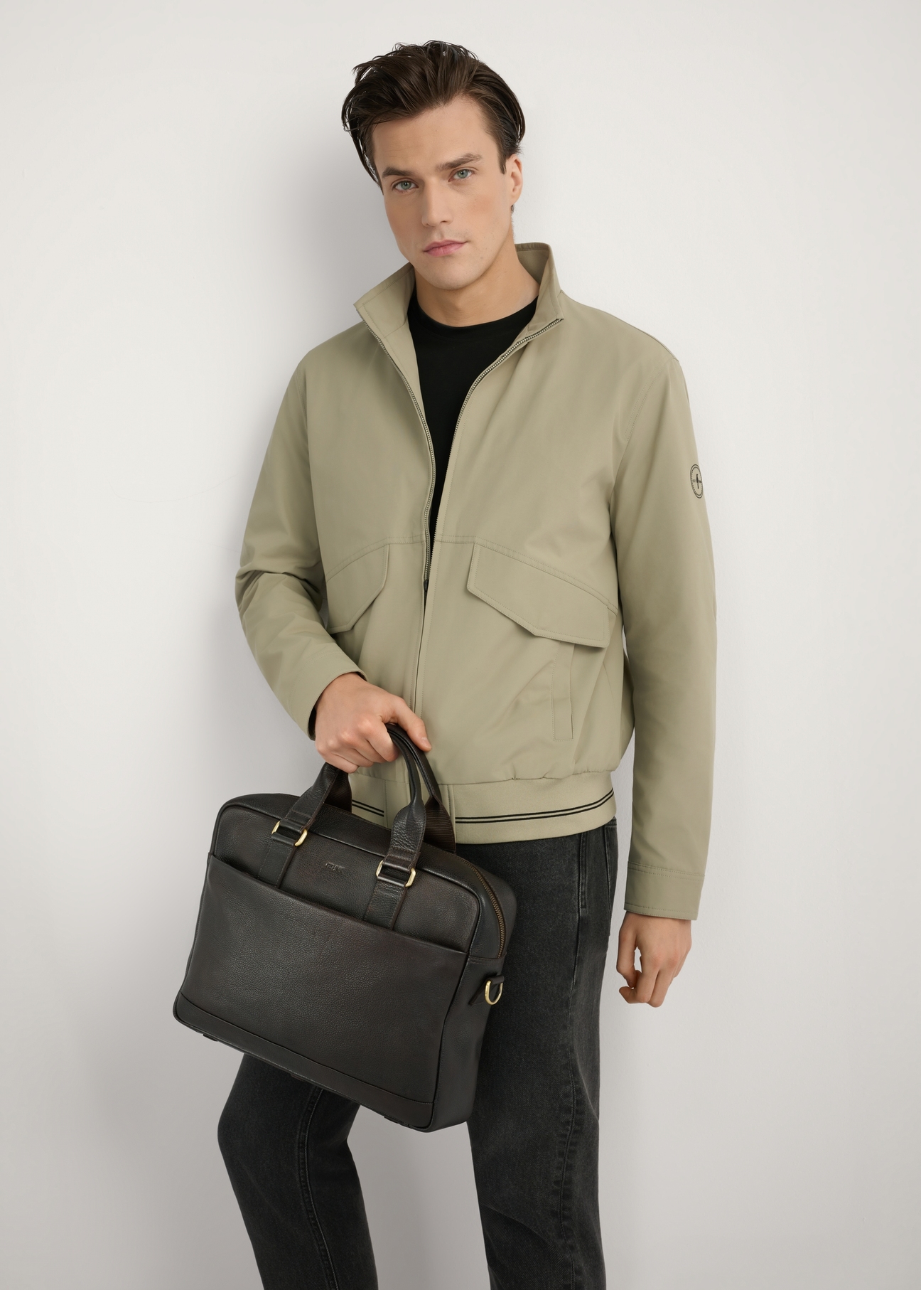 Brązowa skórzana teczka męska TORMS-0503-1I(W26) - widok produktu z przodu, trzymanego przez modela w ręku, na jasnym tle, Widoczna sylwetka do kolan. 