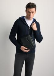 Czarna skórzana saszetka męska TORMS-0251B-99(Z25) - widok produktu z przodu, trzymanego przez modela w ręku, na jasnym tle. Widoczna sylwetka do kolan. 