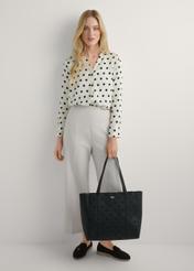 Czarna torebka shopper z monogramem TOREC-1160-9P(W26) - widok produktu, trzymanego przez modelkę w ręku, na jasnym tle. Widoczna cała sylwetka. 