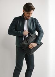 Czarna skórzana torba na laptopa TORMS-0496-99(W26) - widok produktu po skosie, trzymanego przez modela w ręku, na jasnym tle. Widoczna cała sylwetka. 