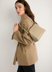 Beżowa torebka damska z monogramem TOREC-1180-1P(W26) - widok produktu lekko po skosie, trzymanego przez modelkę w ręku, na jasnym tle. Widoczna połowa sylwetki.